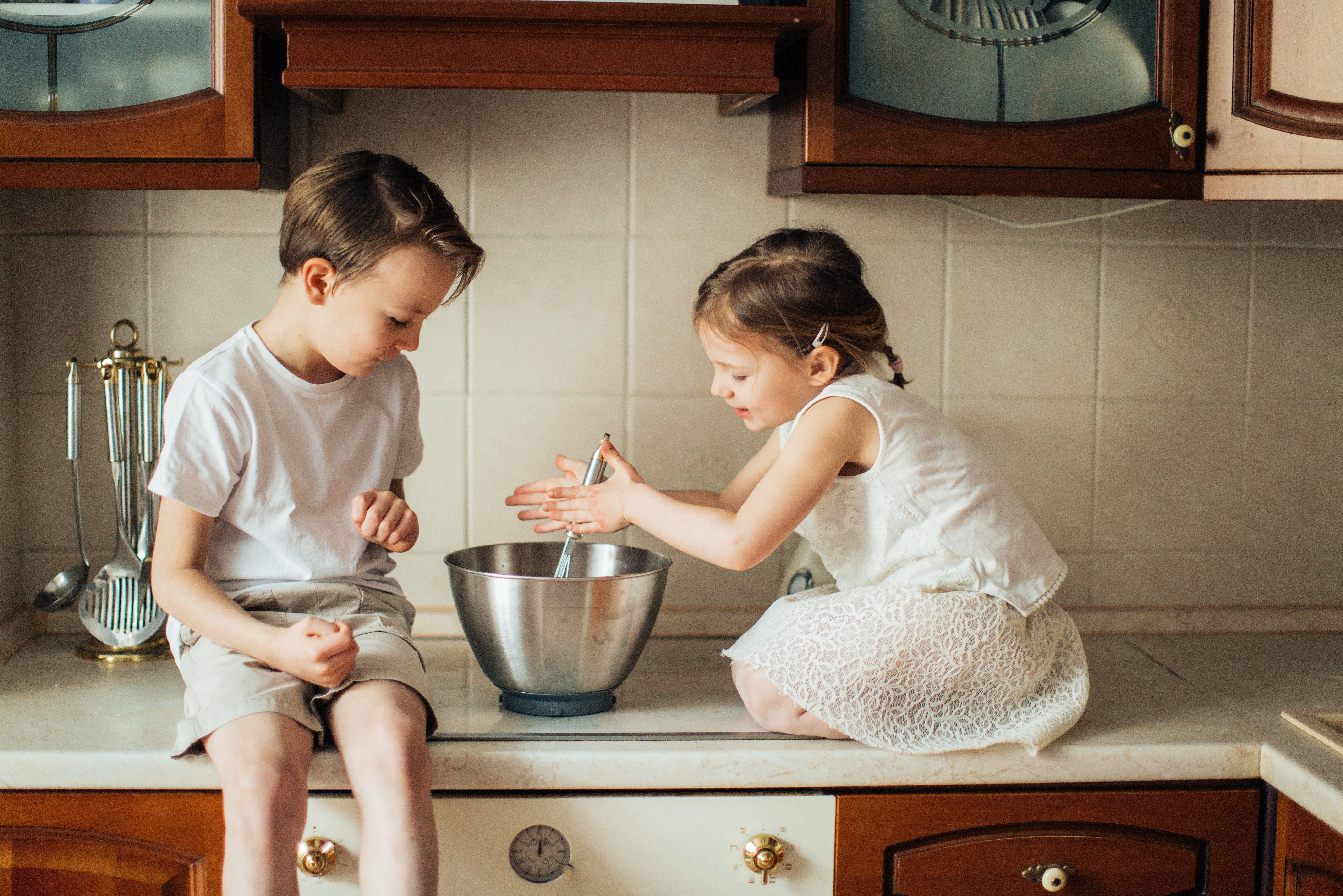 a young boy and a young girl sitting on a kitchen bench with a mixing bowl. 